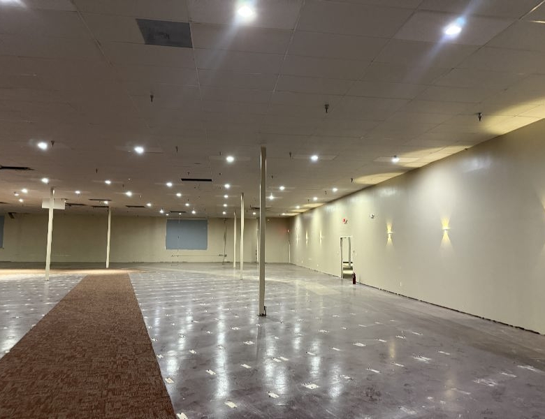 Empty retail space with beige walls, exposed ceiling tiles, and a brown carpet runner down the center.