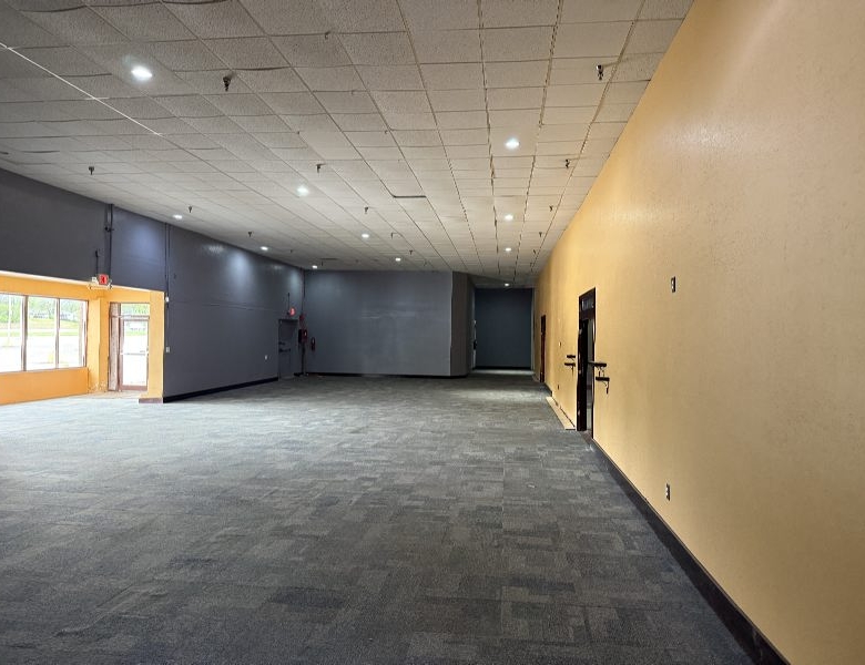 Wide empty hallway in a commercial building with gray left wall, yellow right wall, and overhead fluorescent lights to guide navigation.