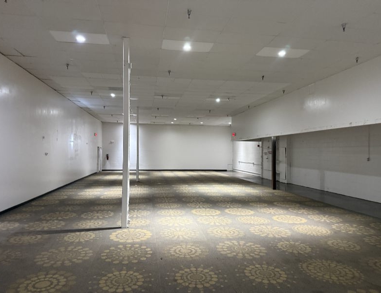 Empty, wide, well-lit room with white walls, a patterned carpet, and a single metal pillar in the center; looks like a vacant event or showroom space.