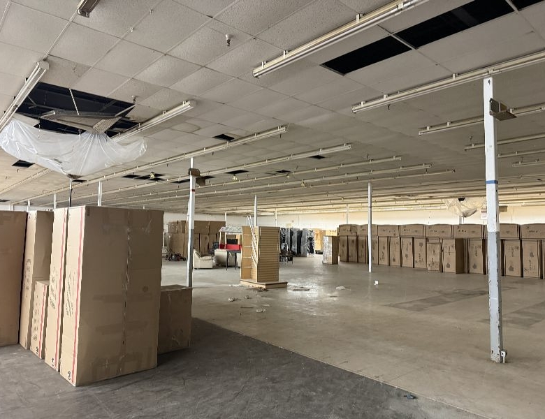 Empty warehouse interior with tall cardboard boxes stacked along the left and far walls, exposed ceiling with missing panels and debris on the floor.
