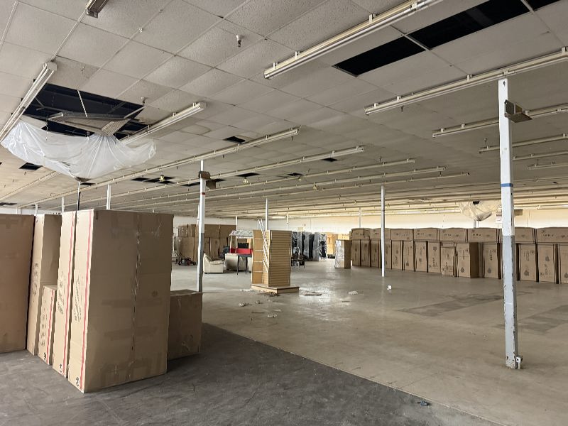 Empty warehouse interior with tall cardboard boxes stacked along the left and far walls, exposed ceiling with missing panels and debris on the floor.