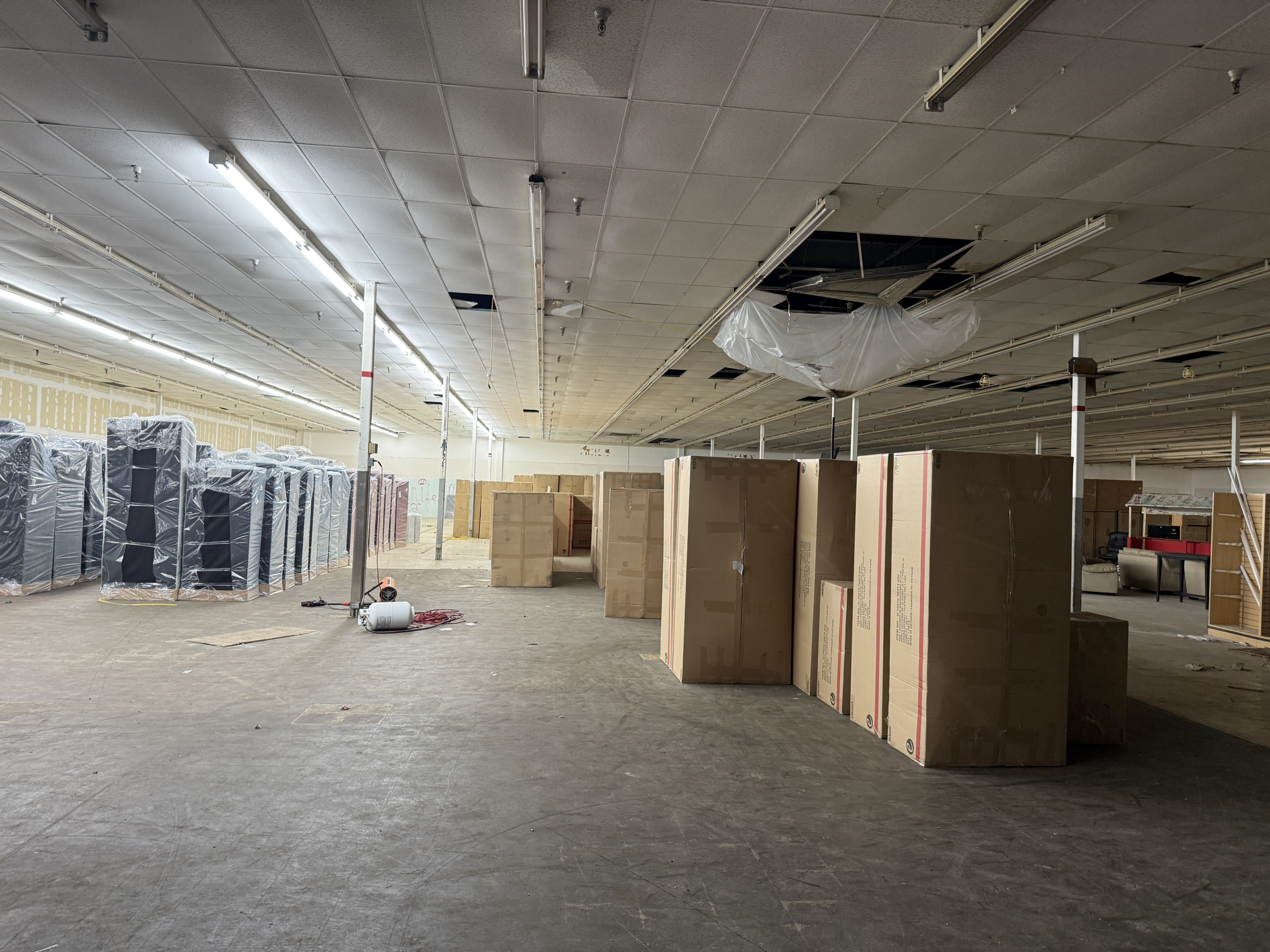 Empty warehouse interior with stacked boxed products and plastic-wrapped pallets along the left, ceiling tiles damaged.