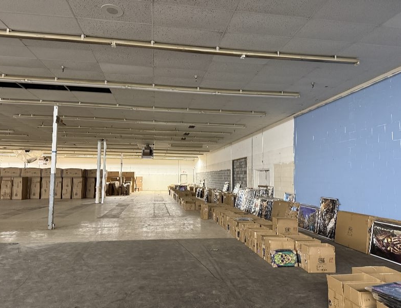 Empty warehouse interior with high ceiling, fluorescent lights, and rows of boxes along the right wall; posters leaned against the boxes.