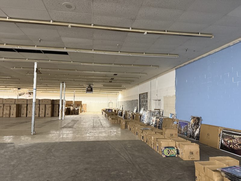 Empty warehouse interior with high ceiling, fluorescent lights, and rows of boxes along the right wall; posters leaned against the boxes.