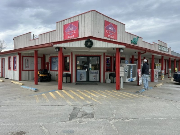 Exterior view of a rustic, single-story storefront with white wooden siding and red trim; a wreath hangs under the awning and a man stands near a vending/payment kiosk outside the entrance.