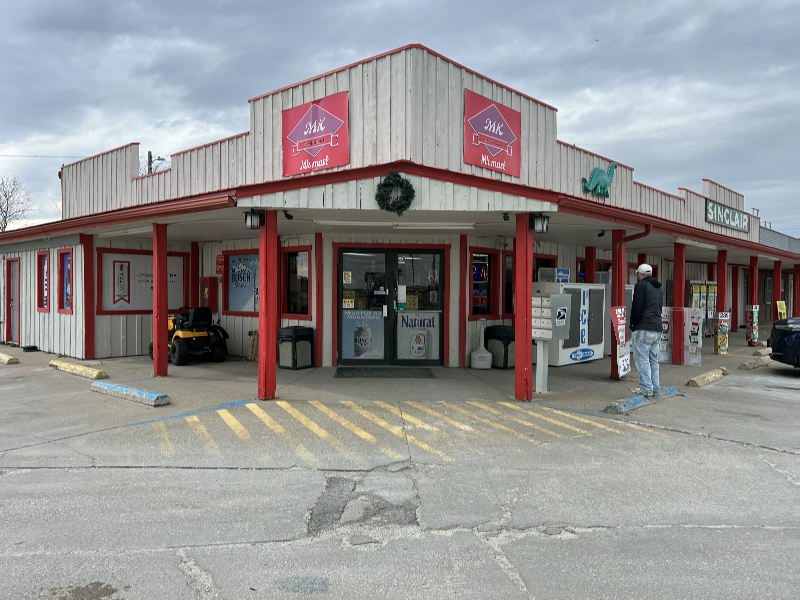 Exterior view of a rustic, single-story storefront with white wooden siding and red trim; a wreath hangs under the awning and a man stands near a vending/payment kiosk outside the entrance.
