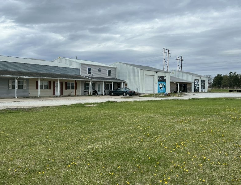 Row of industrial buildings with metal siding along a gravel road, under a cloudy sky; a blue car parked near the entrance.