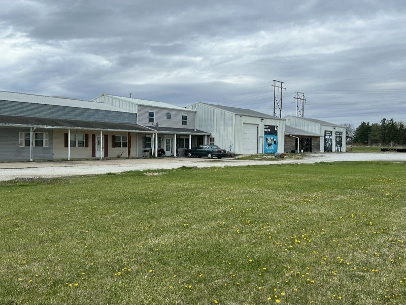 Row of industrial buildings with metal siding along a gravel road, under a cloudy sky; a blue car parked near the entrance.