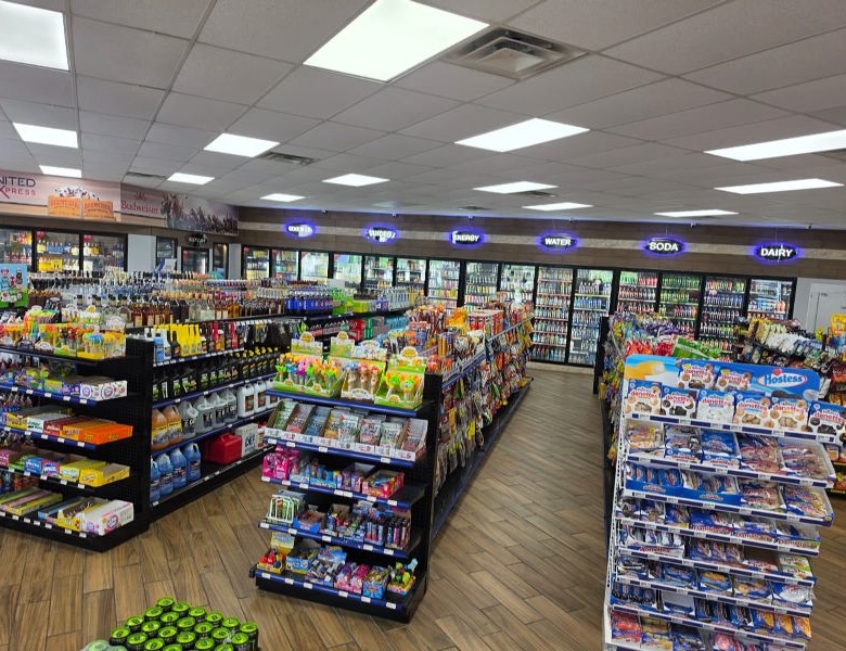 Wide view of a well-lit convenience store with multiple aisles of shelves, colorful snacks, and a row of refrigerated cases along the back wall labeled drinks and dairy.