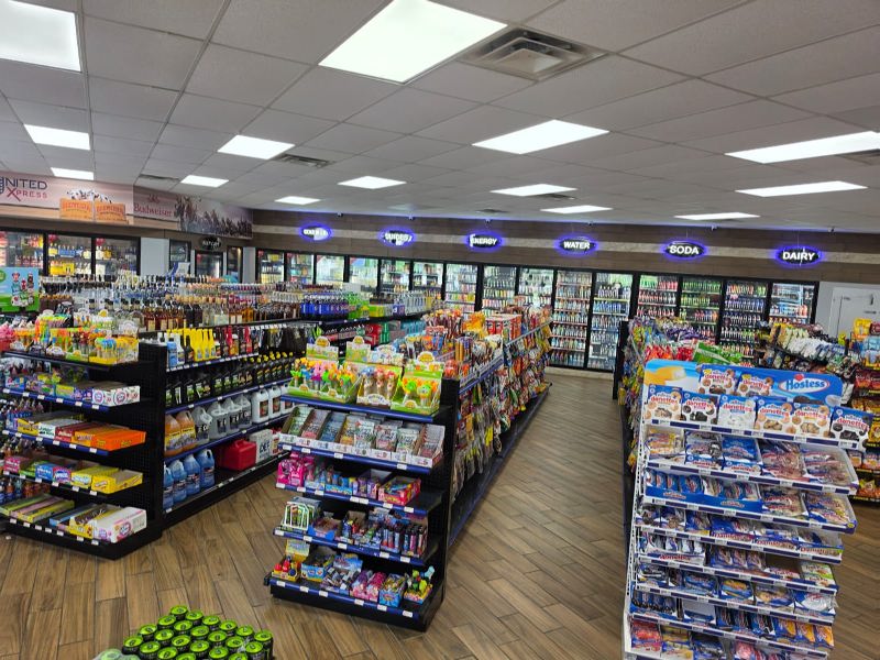 Wide view of a well-lit convenience store with multiple aisles of shelves, colorful snacks, and a row of refrigerated cases along the back wall labeled drinks and dairy.