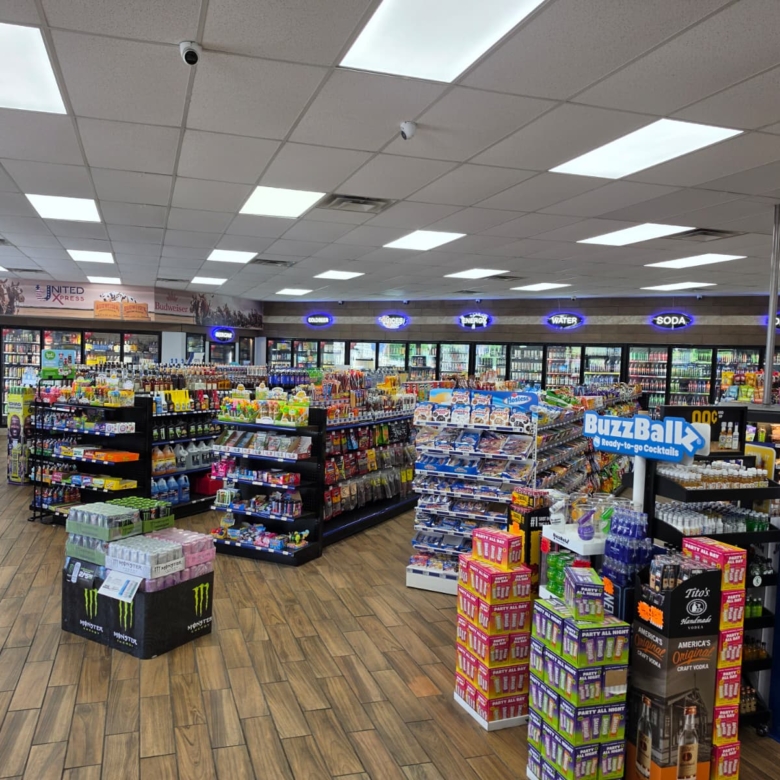Interior of a well-lit convenience store with aisles of snacks and beverages, refrigerated coolers along the back wall, and a large Buzz Ballz display in the foreground.