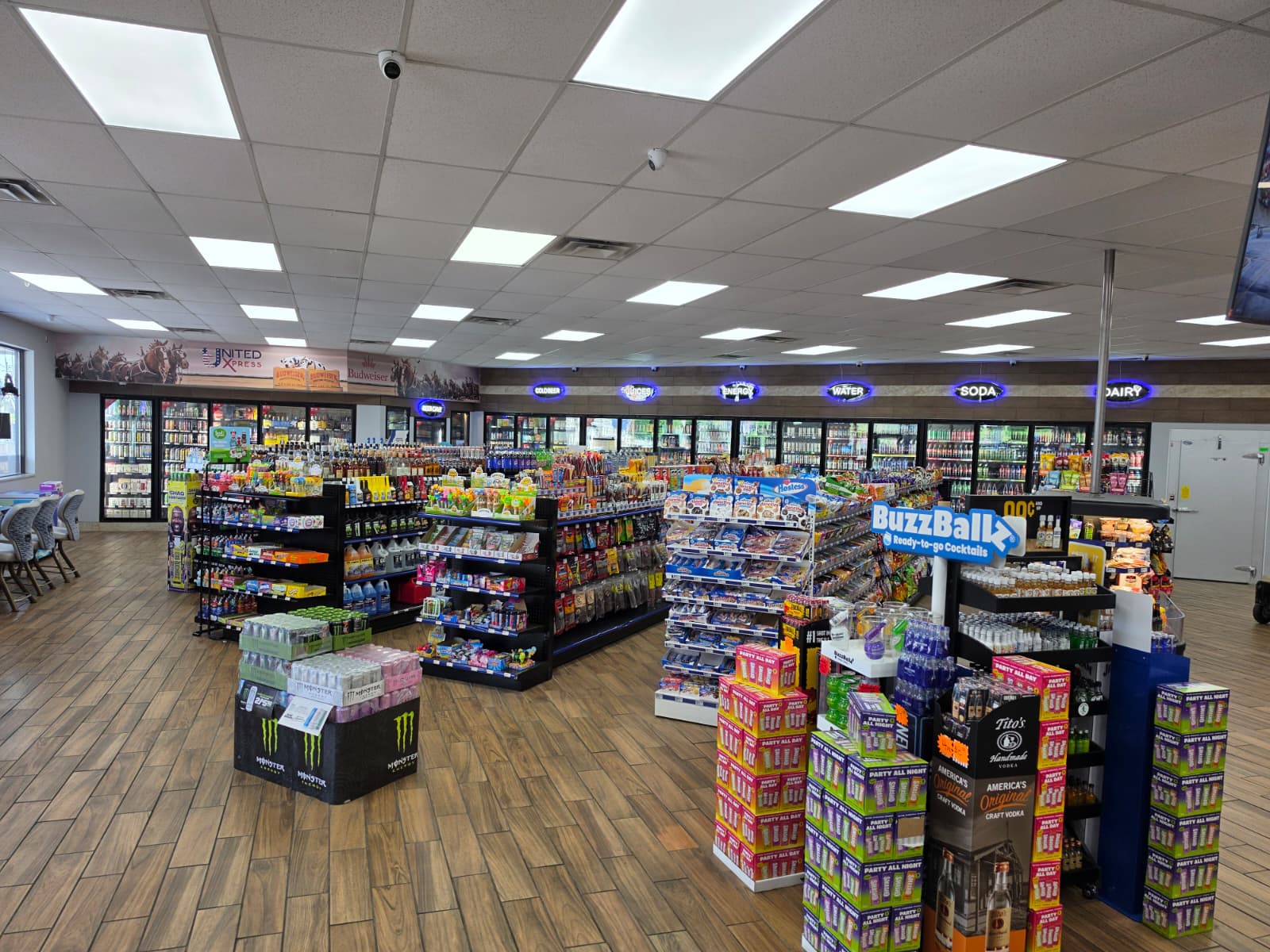 Interior of a well-lit convenience store with aisles of snacks and beverages, refrigerated coolers along the back wall, and a large Buzz Ballz display in the foreground.