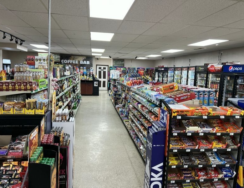 Convenience-store aisle with snack racks on both sides, refrigerated drinks on the right, and a coffee bar area visible in the back.