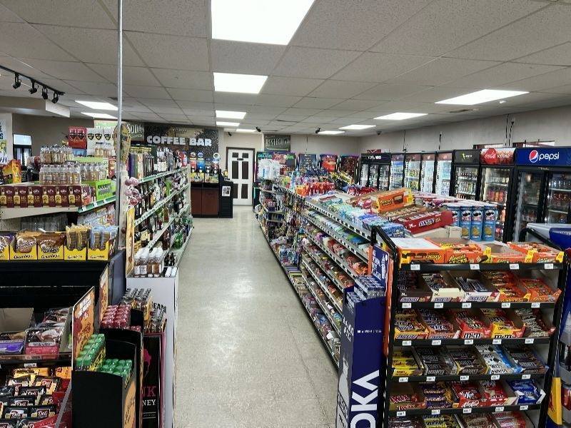 Convenience-store aisle with snack racks on both sides, refrigerated drinks on the right, and a coffee bar area visible in the back.