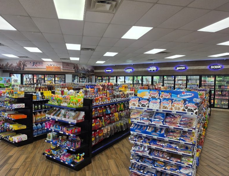 Bright grocery store aisle with shelves of snacks, candy, and drinks, and refrigerated coolers along the back wall.