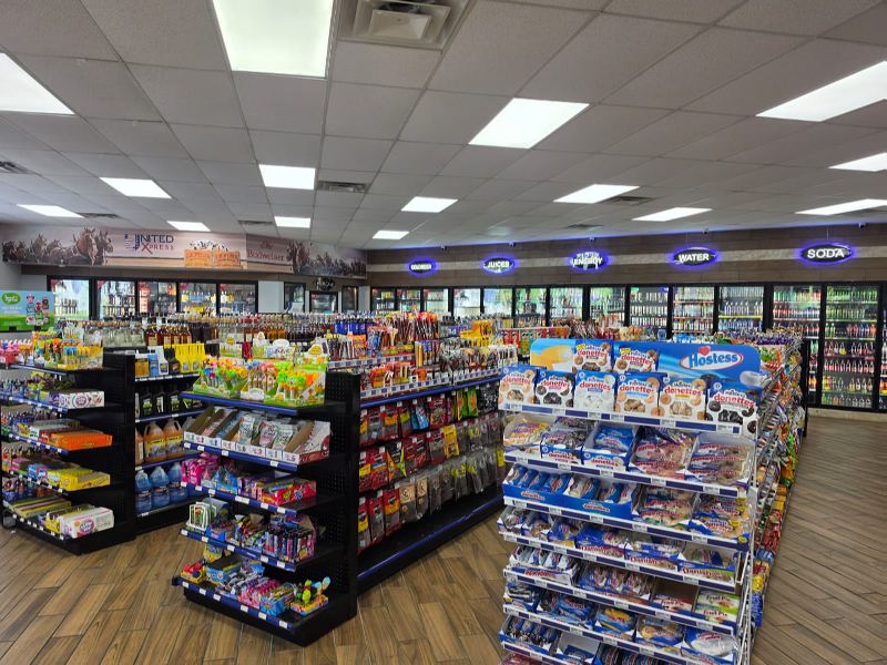 Bright grocery store aisle with shelves of snacks, candy, and drinks, and refrigerated coolers along the back wall.