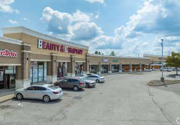 Outdoor strip mall with multiple storefronts, including Beauty & Company, and several parked cars under a blue, partly cloudy sky.