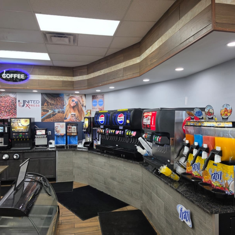 Cafe counter with self-serve beverage machines, including Pepsi and Coca-Cola taps, and juice dispensers under a neon COFFEE sign.