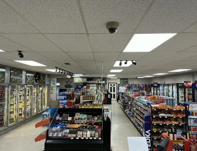 Bright grocery store interior with a long central aisle, refrigerated displays along the left wall, and snack aisles on the right; ceiling grid and security camera visible.