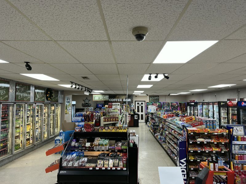 Bright grocery store interior with a long central aisle, refrigerated displays along the left wall, and snack aisles on the right; ceiling grid and security camera visible.