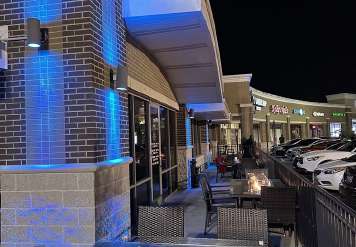 Nighttime view of a brick storefront with blue neon lights, outdoor seating, and parked cars along a strip mall.”,