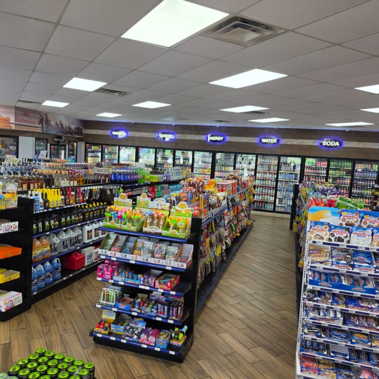 Supermarket aisle with long displays of snacks, drinks, and cleaning supplies; refrigerated sections at the back under bright ceiling lights.