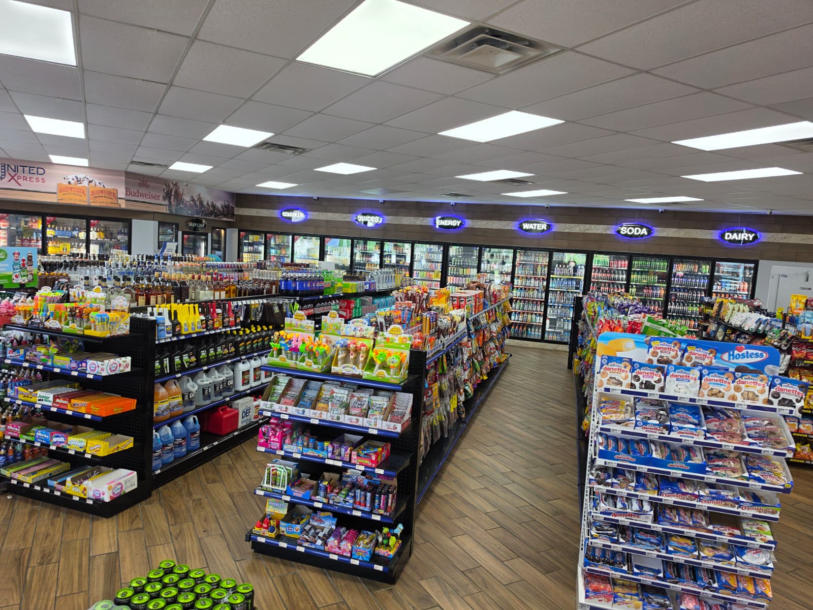 Supermarket aisle with long displays of snacks, drinks, and cleaning supplies; refrigerated sections at the back under bright ceiling lights.