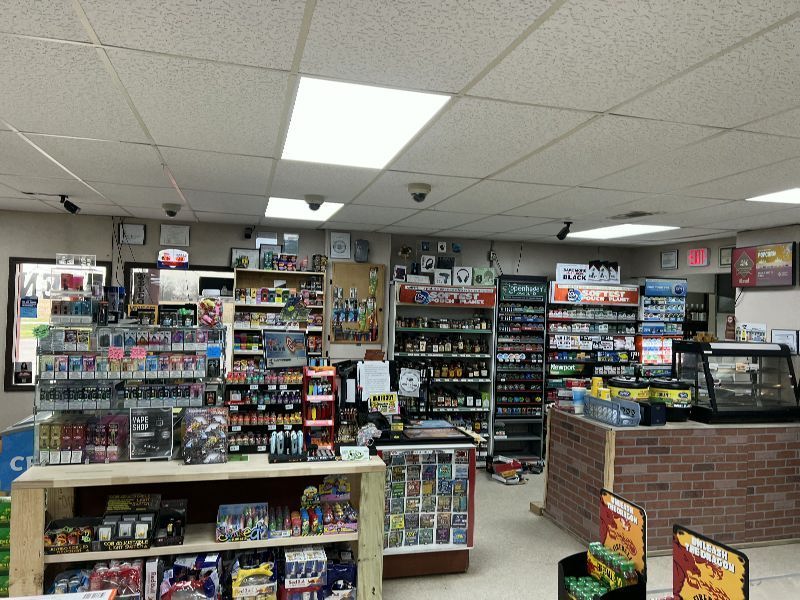 Interior of a small convenience store with shelves full of snacks, drinks, and assorted items; a brick-patterned checkout counter is on the right."