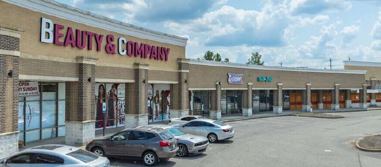 Wide view of a shopping plaza with a prominent 'Beauty & Company' store and other storefronts, parked cars in front, under a cloudy sky.