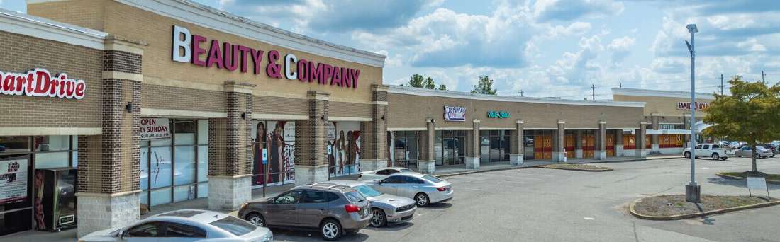 Wide view of a shopping plaza with a prominent 'Beauty & Company' store and other storefronts, parked cars in front, under a cloudy sky.