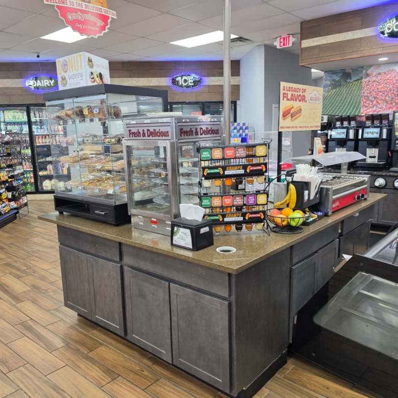 Interior of a convenience store with a central service counter and glass display case of baked goods; snacks, drinks, and coffee machines visible in background.
