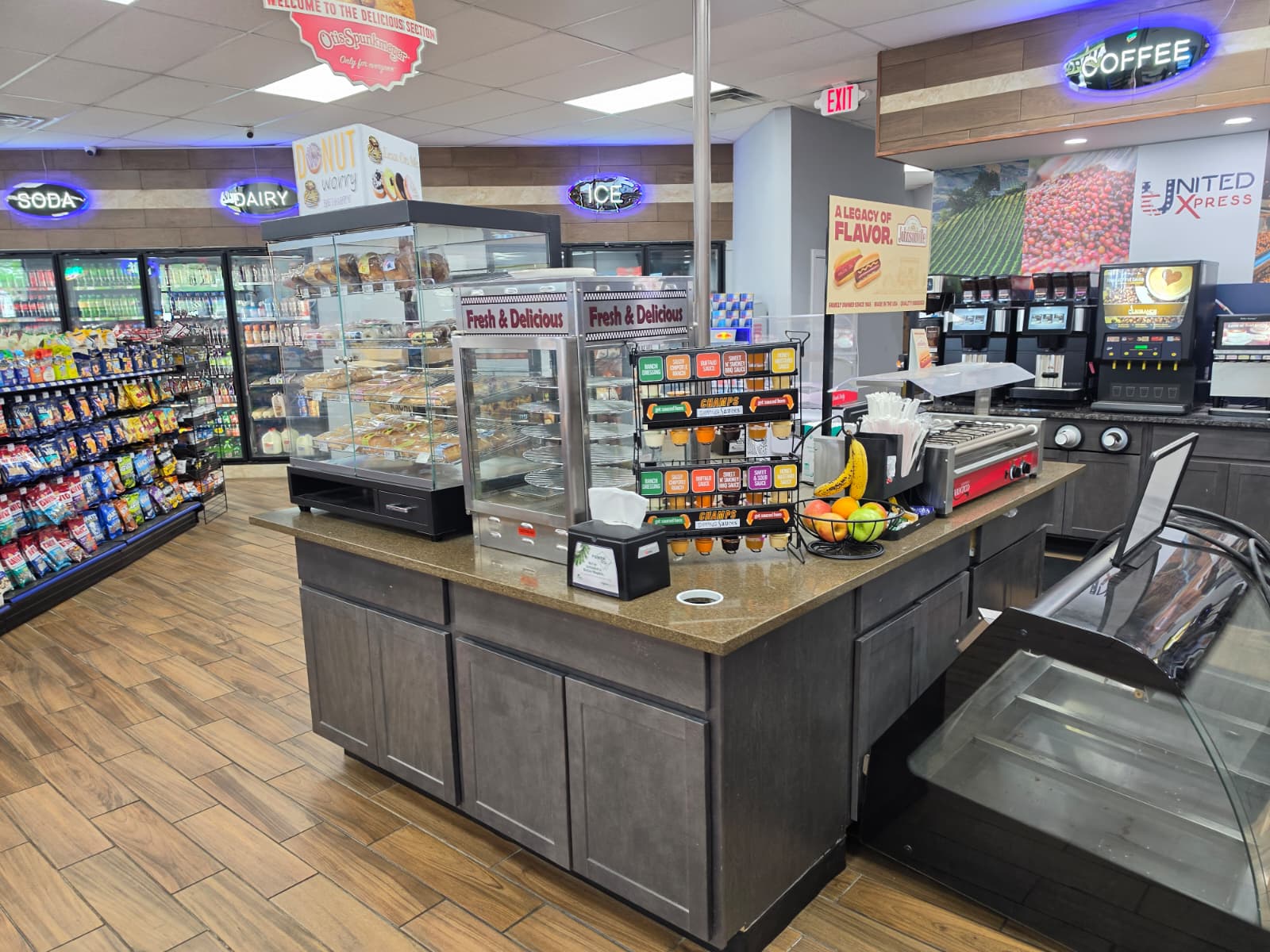 Interior of a convenience store with a central service counter and glass display case of baked goods; snacks, drinks, and coffee machines visible in background.