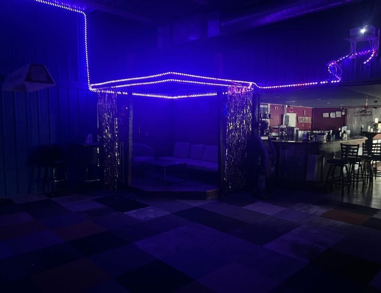 Neon-lit lounge area with a blue-purple gazebo-like booth and shimmering curtains under purple lights, bar area to the right.