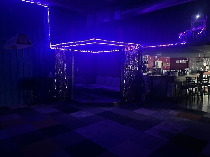 Neon-lit lounge area with a blue-purple gazebo-like booth and shimmering curtains under purple lights, bar area to the right.