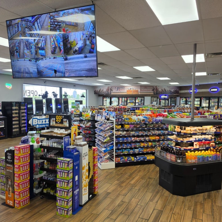 Interior of a bright convenience store with snack displays, beverage coolers, and a large overhead multi-panel security monitor showing multiple camera angles (CCTV).