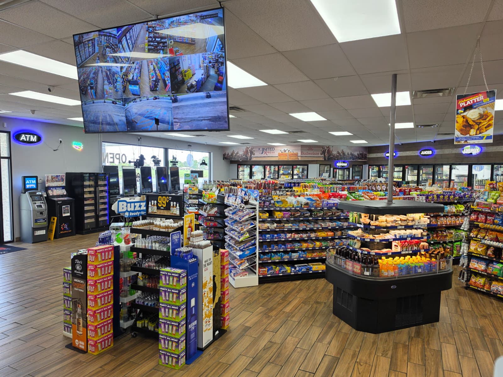 Interior of a bright convenience store with snack displays, beverage coolers, and a large overhead multi-panel security monitor showing multiple camera angles (CCTV).
