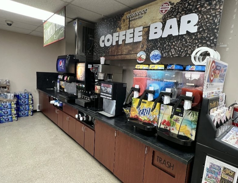 Interior of a convenience-store coffee bar with beverage dispensers, a coffee machine, and a trash bin under the counter; large 'COFFEE BAR' sign on the wall