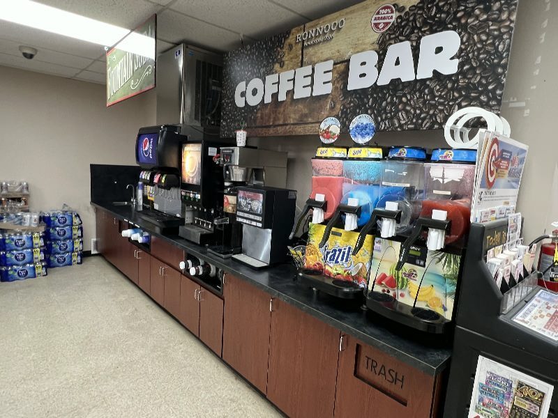 Interior of a convenience-store coffee bar with beverage dispensers, a coffee machine, and a trash bin under the counter; large 'COFFEE BAR' sign on the wall
