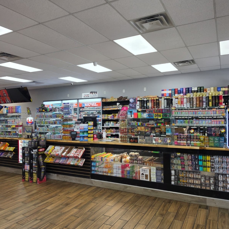 Wide view of a well-lit convenience store with long shelves of snacks, drinks, and tobacco products along a back wall, and a wooden floor.
