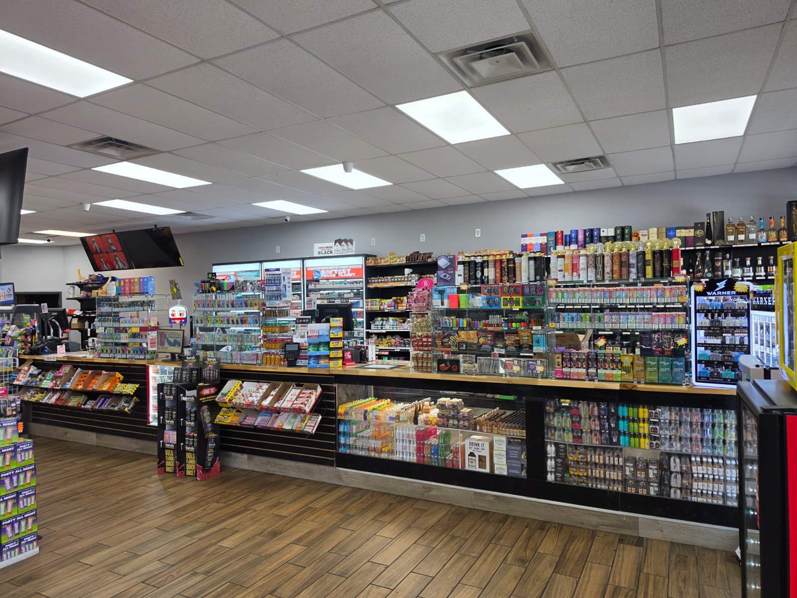 Wide view of a well-lit convenience store with long shelves of snacks, drinks, and tobacco products along a back wall, and a wooden floor.