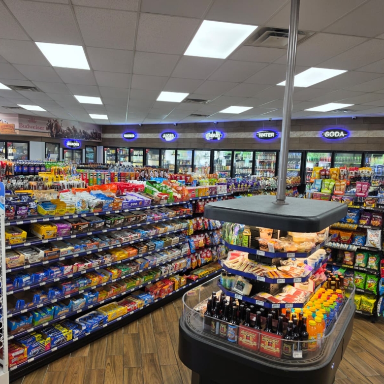 A well-stocked convenience store aisle with snack shelves, coolers in the back, and neon signs along the wall above the refrigerated section.