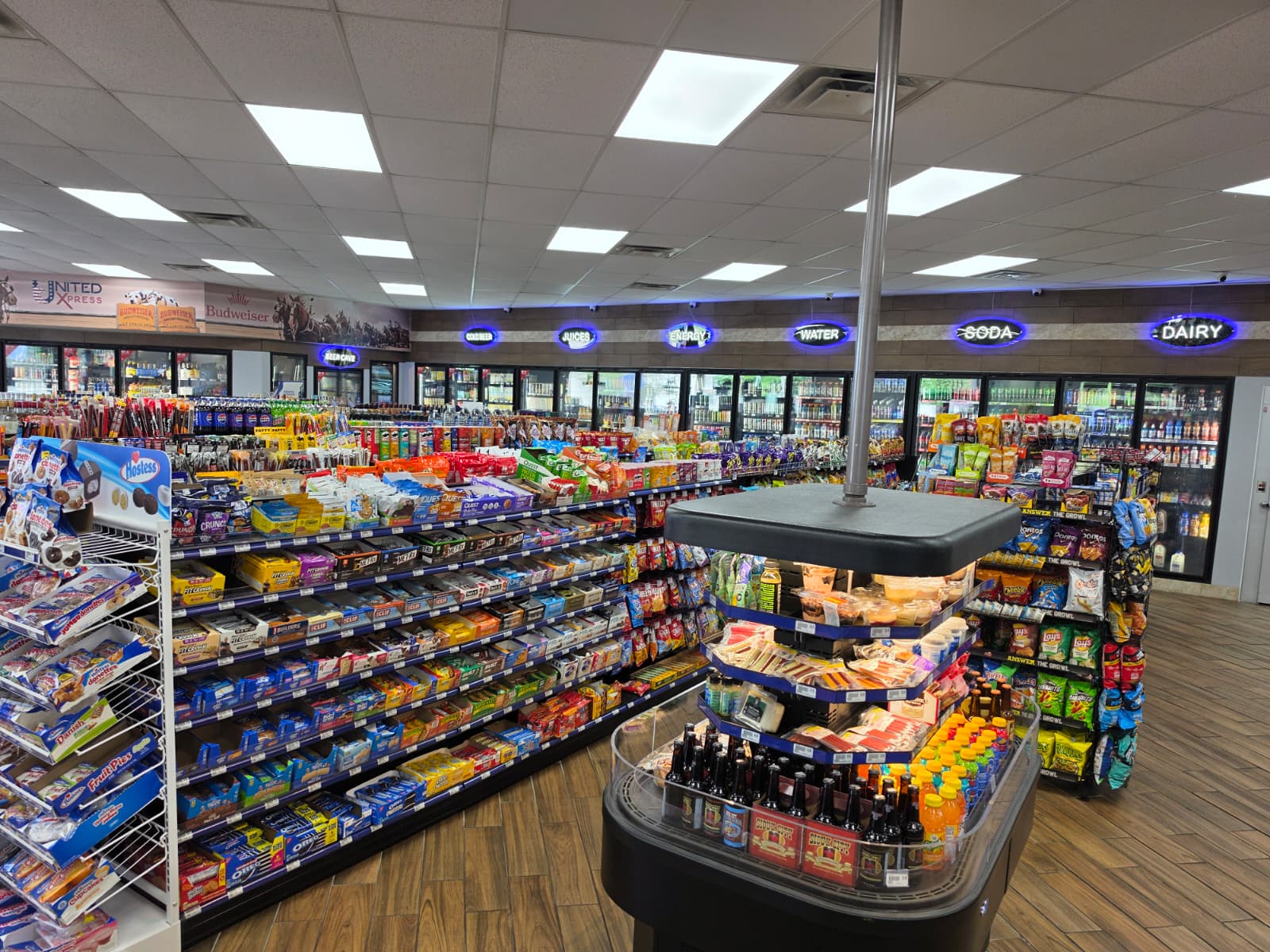 A well-stocked convenience store aisle with snack shelves, coolers in the back, and neon signs along the wall above the refrigerated section.