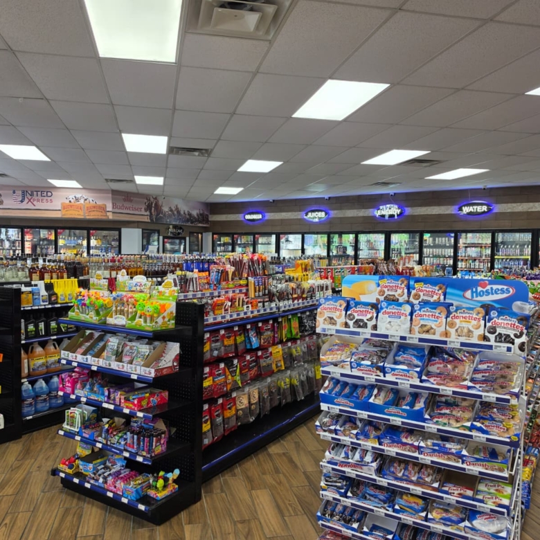 Bright store interior with multiple shelves of snacks in the foreground and refrigerated drinks along the back wall.