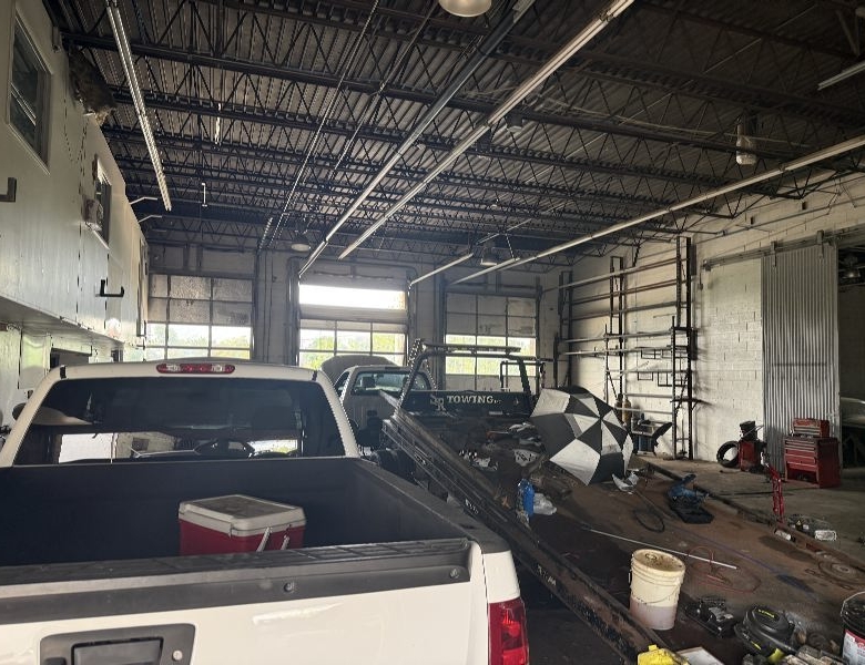 Auto repair shop scene with a white pickup bed in the foreground and tools, ladders, and vehicles inside a large garage bay.