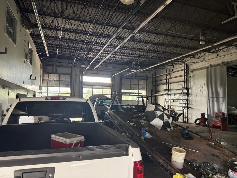Auto repair shop scene with a white pickup bed in the foreground and tools, ladders, and vehicles inside a large garage bay.