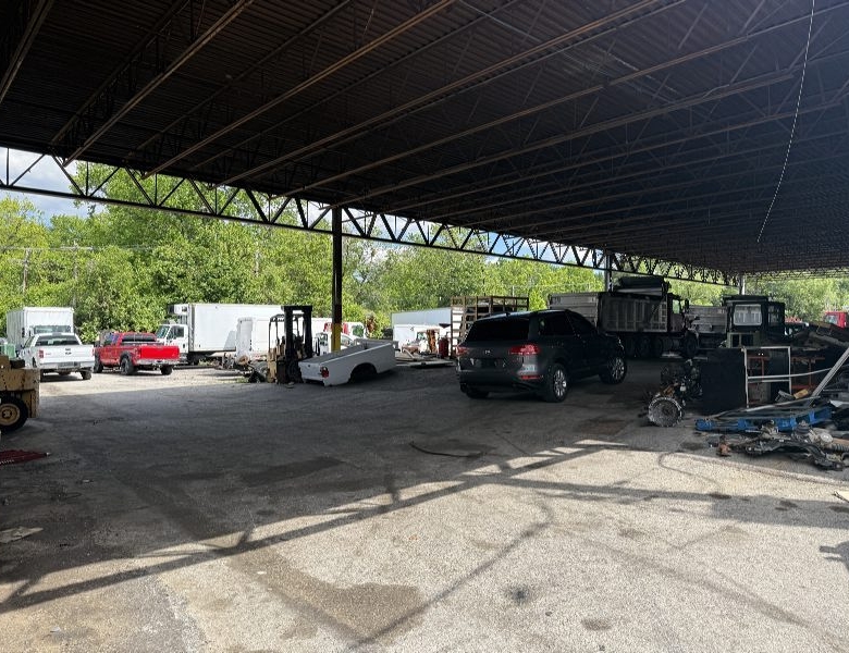 Open-air salvage yard under a corrugated metal roof with vehicles, forklifts, and scattered equipment in a dusty lot.