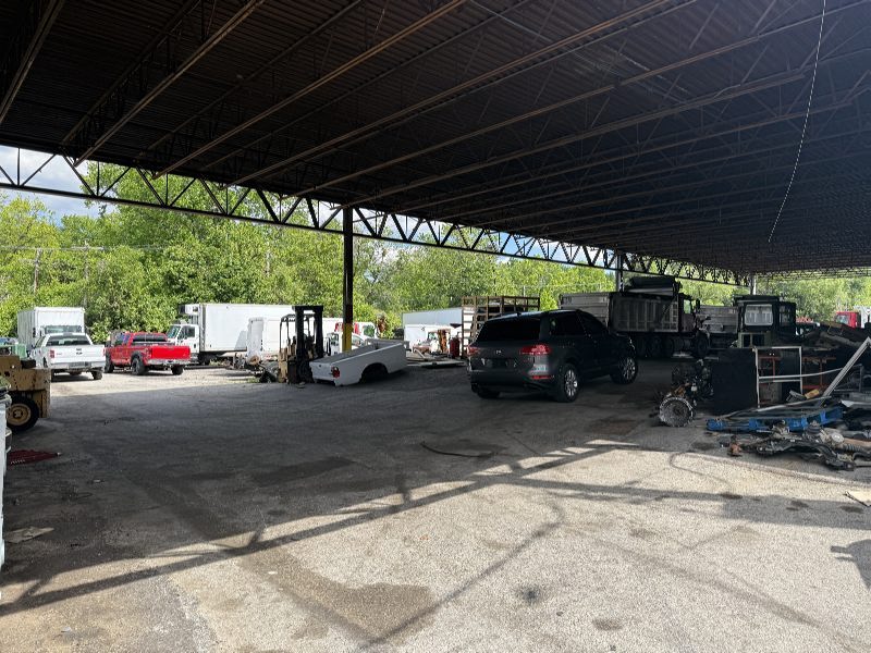 Open-air salvage yard under a corrugated metal roof with vehicles, forklifts, and scattered equipment in a dusty lot.