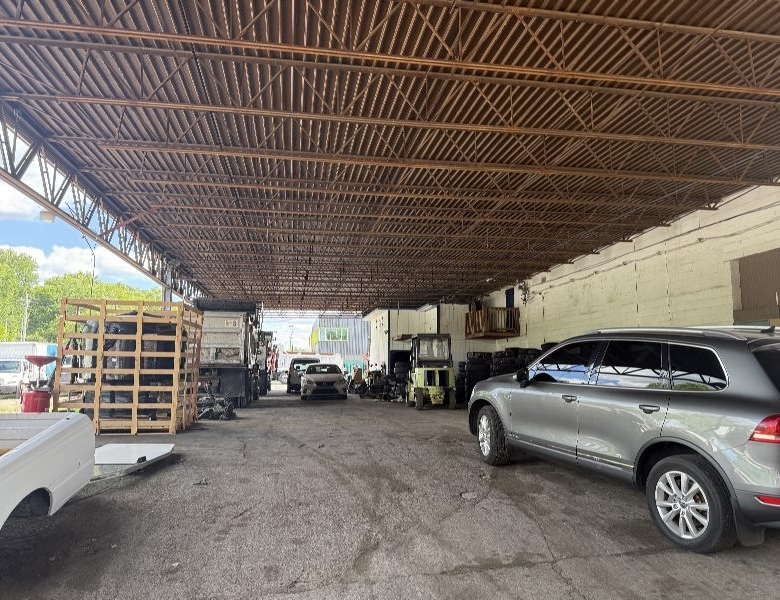 A large open-sided workshop with a metal truss roof, several cars parked inside, forklift and pallets along the left wall under a high ceiling.