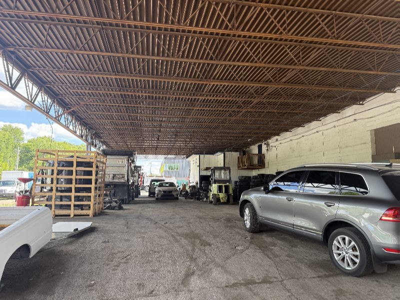 A large open-sided workshop with a metal truss roof, several cars parked inside, forklift and pallets along the left wall under a high ceiling.