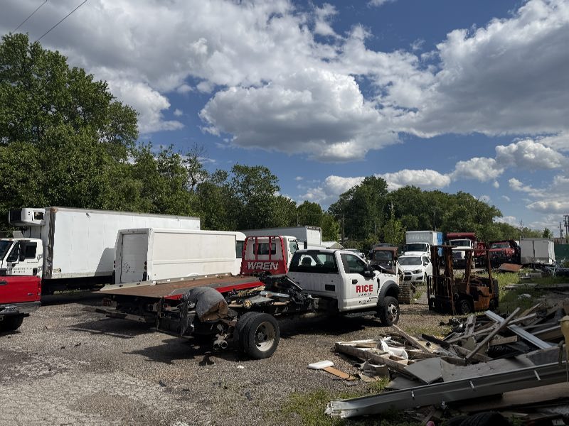 Outdoor junkyard with several white and red trucks, a flatbed trailer, and scattered metal scrap under a blue sky with clouds.